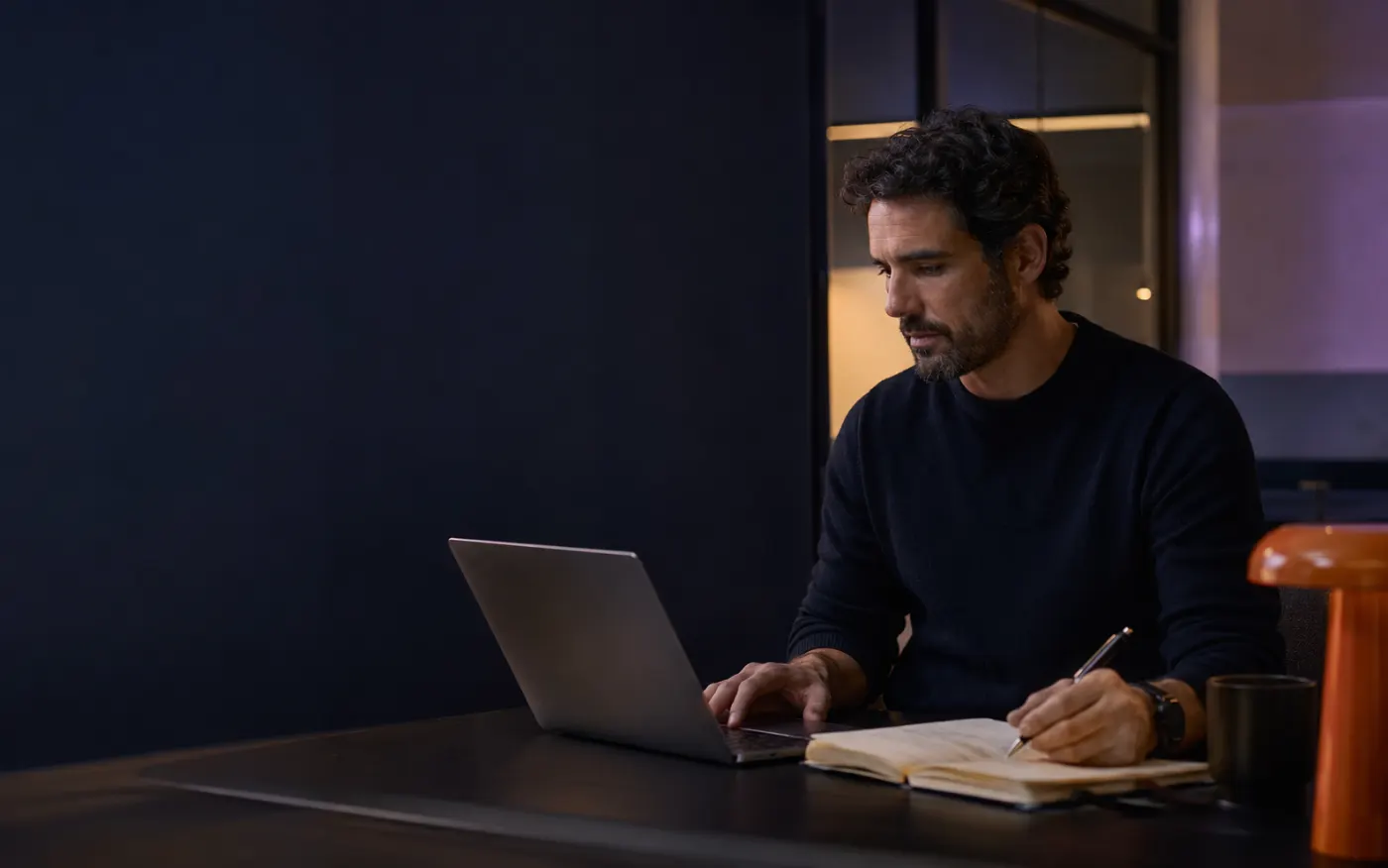 Persona trabajando de noche en un escritorio, tomando notas junto al portátil
